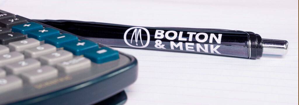 Close-up of a calculator with black buttons on the left and a black pen featuring the Bolton & Menk logo on the right, resting on lined paper, symbolizing meticulous calculations for project funding support.
