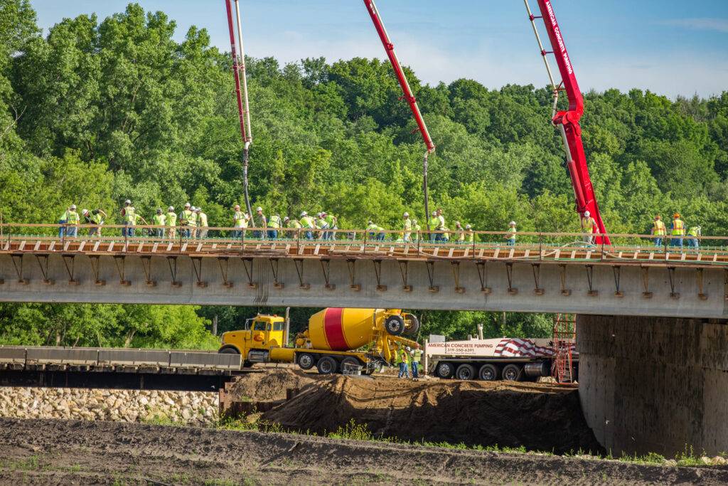 Amidst the backdrop of greenery and clear blue skies, construction workers diligently offer structural services on a bridge. Two large red concrete pumps are in use, while a cement mixer and truck are stationed below, ready to support their efforts.