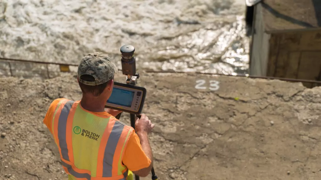 A person in an orange safety vest and camouflage hat engages in land surveying near a body of water. They use equipment mounted on a tripod, with a tablet facing the water currents below. Numbers 2 3 are painted on the concrete.