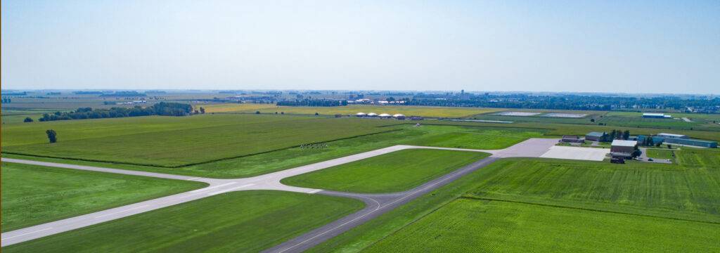 Aerial view of a small rural airport surrounded by expansive green fields. The runway and taxiways, integral to its aviation services, are visible alongside a few hangars and buildings. The landscape stretches into the distance under a clear blue sky.