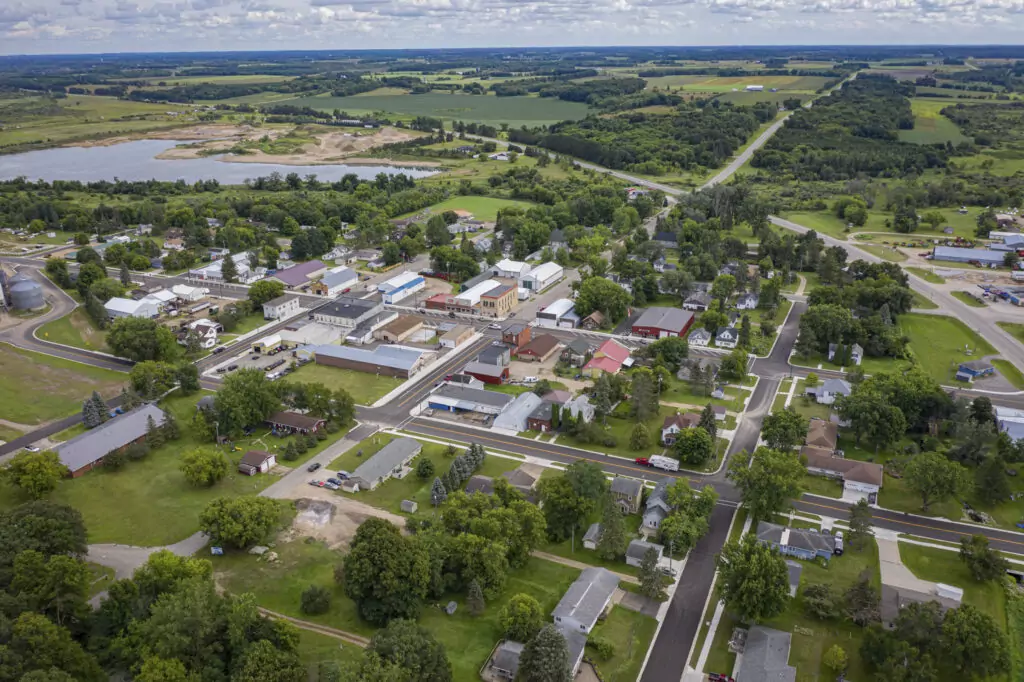 Aerial view of a small town reveals the meticulous art of civil engineering with its grid of streets, houses, and trees. A body of water graces the left side, embraced by fields and greenery. The horizon unfolds more fields and forest patches under a partly cloudy sky.
