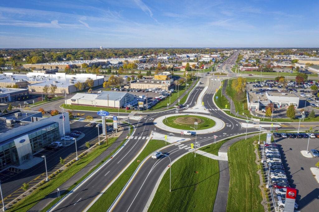 From an aerial view, witness the intricacies of transportation engineering as a circular roundabout efficiently channels vehicles between suburban roads. A car dealership stands in the foreground, with residential and commercial buildings under a clear blue sky completing the scene.
