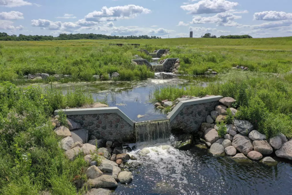 A lush green meadow with a stone-lined stream featuring small cascading waterfalls showcases the harmony of nature and Water Resources Engineering. The scene, dotted with wild grass and rocks under a partly cloudy sky, reveals a wooden bench and silo in the background.