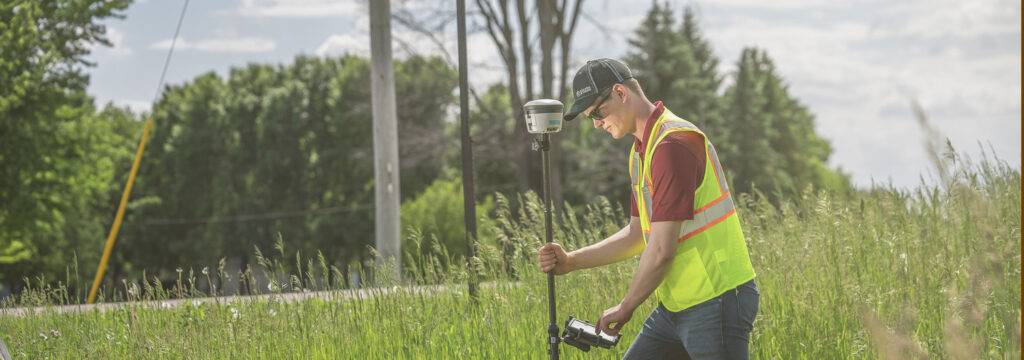 A person in a neon safety vest and cap uses a GPS survey tool in a grassy field, integrating Geographic Information Systems. They are crouched slightly, holding the tripod-mounted device with one hand. Trees, a utility pole, and a cloudy sky form the backdrop.