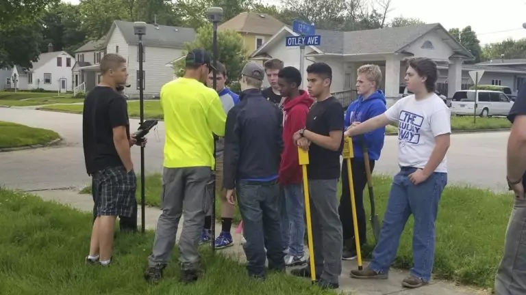 A group of young people and two adults from a professional services firm stand on a grassy sidewalk corner near a street sign that reads Otley Ave. Some hold surveying tools, and one adult in a yellow shirt appears to be instructing them. Residential houses are in the background.