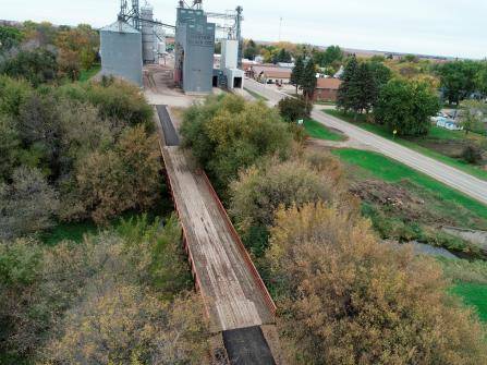 Aerial view of a wooden bridge surrounded by trees with autumn foliage. In the background, there are large industrial silos, a small road, and some buildings. The sky is overcast, giving the scene a muted, serene atmosphere.