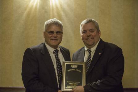 Two men in suits stand side by side, smiling. The man on the right holds a plaque with the title Founder Award. The background features a softly lit, warm-toned wall.