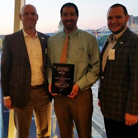 Three men stand together in front of a window, with one man holding a plaque. They are dressed in business attire, and the cityscape is visible outside the window.