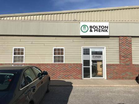 A single-story building with a Bolton & Menk sign above the door. There are two windows and a brick and siding facade. A parked car is partially visible in the foreground.