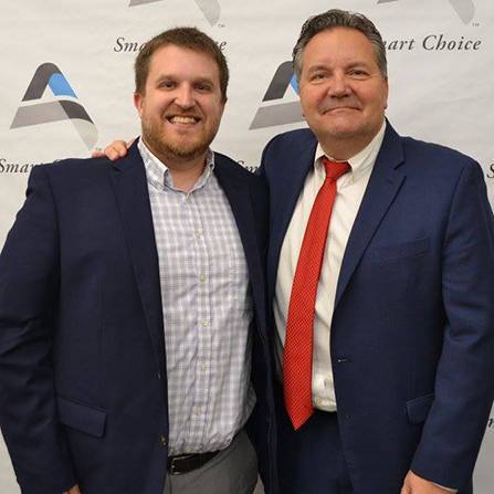 Two men in suits are smiling at the camera. The man on the left wears a checked shirt and blazer, while the man on the right sports a red tie. They are standing in front of a backdrop with the words Smart Choice and a stylized logo.