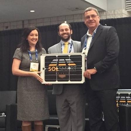 Three people stand together smiling, holding a large box with a solar panel design and SOX SMART on it. The woman on the left is wearing a gray dress, the man in the middle has on a suit and tie, and the man on the right is in a dark suit.