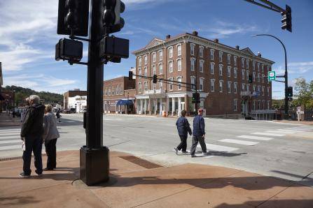 A street corner with two people crossing a crosswalk. In the background, theres a large, three-story brick building with multiple windows. The sky is clear with scattered clouds, and a traffic light stands to the left.