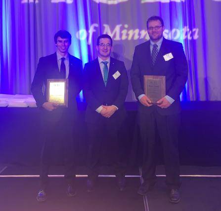 Three men in suits stand on stage holding plaques. The backdrop features blue lighting and the partial text of Minnesota.