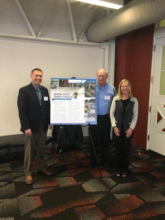 Three people stand in a room next to an easel with a poster titled Buffalo Tract & Joe Stacey Parkland. The two men and one woman smile at the camera. The room has white walls, a red accent wall, and a closed door.