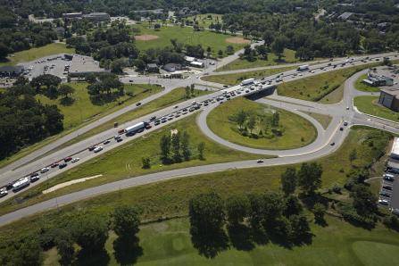 Aerial view of a highway interchange with multiple looping ramps. Vehicles are lined up, causing traffic congestion on one side. Surrounding areas include green spaces, trees, a parking lot, and open fields.