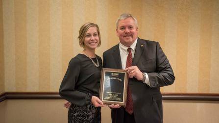 Two people stand indoors holding an award plaque together. The woman on the left is wearing a black outfit, and the man on the right is in a suit with a red tie. They are smiling in front of a beige, striped wall.