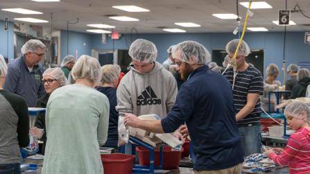 A group of people wearing hairnets work together in an indoor setting, possibly a food packaging area. They are handling items on tables and using equipment, engaged in a collective task. The room has overhead lighting and visible equipment.