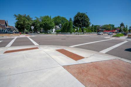 A street intersection with crosswalks is visible under a clear blue sky. Trees and houses line the streets in the background. The sidewalks have tactile paving for accessibility.