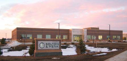 A modern brick building is pictured at sunset with a pink sky. A sign in the foreground reads Bolton & Menk. The ground is partially covered in snow, and small evergreen trees are planted near the entrance.