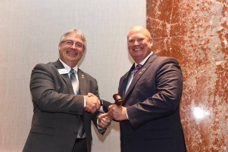 Two men in suits shake hands while one hands a gavel to the other. They stand in front of a beige and red marble wall, smiling for the camera.