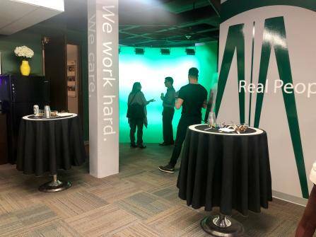 Office lounge with small tables covered in black tablecloths, holding drinks and snacks. Four people stand in conversation near a bright green backdrop. Walls display motivational phrases, and there is a water cooler nearby.