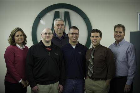 Six people standing in front of a wall with a circular logo. Three men are wearing glasses. Two women are present, one wearing a red sweater. They all stand close together, smiling, in a casual office environment.