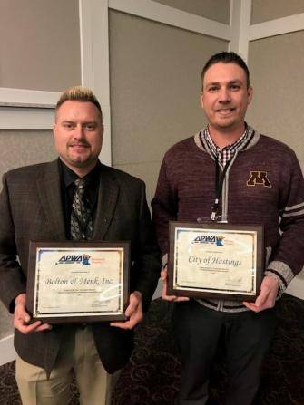 Two men are standing indoors, each holding an award plaque. The man on the left is wearing a dark suit, while the man on the right is in a burgundy sweater with an M logo. The plaques have text but are not fully readable in the image.