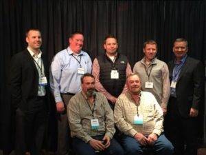 Seven men are posing for a group photo in front of a black curtain. Four are standing in the back, and three are seated in front. They are wearing a mix of business and casual attire, each with a name badge.