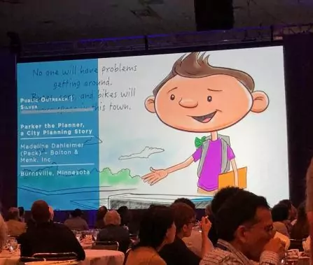 A conference room with attendees facing a large screen displaying a cartoon character named Parker the Planner. The slide includes text about public outreach in Burnsville, Minnesota.