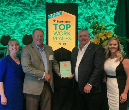 Four people standing together, smiling, holding an award plaque in front of a background with a Star Tribune Top Workplaces 2019 banner and green foliage.