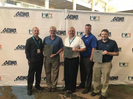 Five men standing in front of a backdrop with APWA and PWX Public Works Expo logos, holding plaques. They are smiling and dressed in business casual attire. The setting appears to be an indoor event or conference.