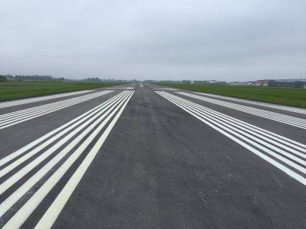 A wide, empty airport runway stretching into the distance under a cloudy sky. The runway is lined with white markings, surrounded by green grass on both sides, and buildings are visible in the far background.