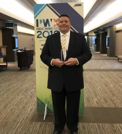 A man in a suit holds a trophy while standing in front of a large sign that reads PWX Public Works Expo 2019. The setting appears to be a hallway or lobby area.