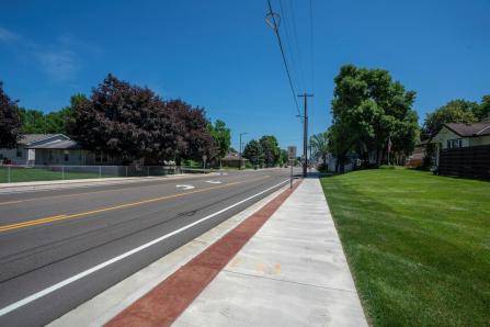 A suburban street with a wide sidewalk, bordered by green lawns and trees. Houses are visible on the left side, under a clear blue sky. Power lines run alongside the road.