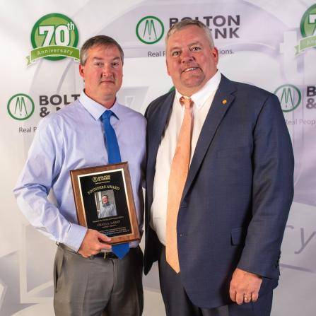 Two men in formal attire pose for a photo. One holds a plaque. Behind them is a backdrop featuring the Bolton & Menk logo and a 70th Anniversary emblem.