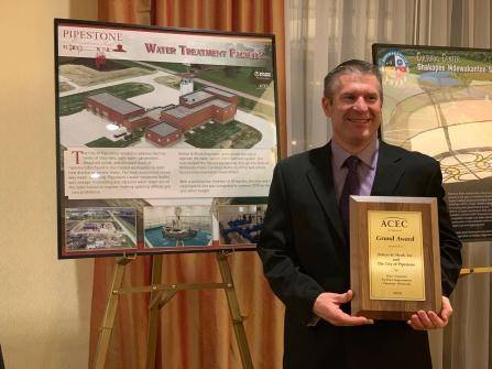 A person in a suit holds a plaque labeled ACEC Grand Award. Behind them, there are two posters, one depicting a water treatment facility and the other related to water systems.