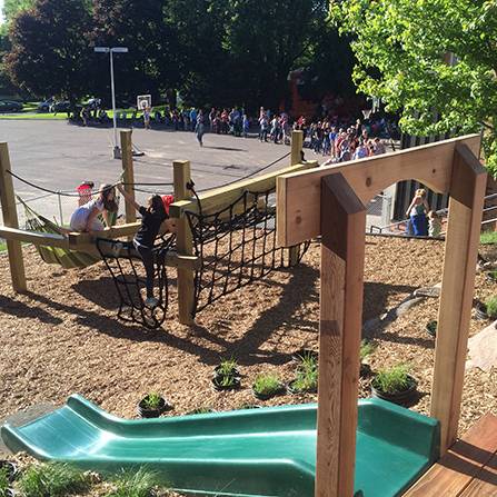 Children playing on a playground with a slide, climbing net, and wooden structures. A group of people is gathered in the background on a paved area. The scene is sunny with trees surrounding the area.
