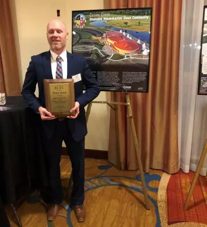 A man in a suit holds an award plaque, standing in front of a poster about the Shakopee Mdewakanton Sioux Community project. The setting is indoors with curtains and carpet visible in the background.