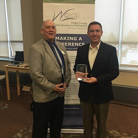 Two men standing indoors, one holding a glass award. Both are wearing suits. Behind them is a banner with Wright County Economic Development Partnership written on it. Theres a table and a window in the background.