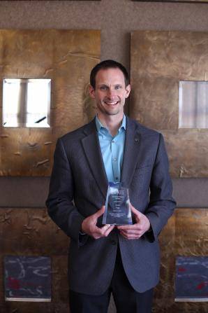 A person in a blue shirt and gray blazer is holding an award. They are standing in front of a textured, gold-colored wall with square art pieces.