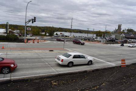 A busy intersection with multiple cars on a cloudy day. Orange traffic cones and barricades are positioned along the road. Buildings and trees are visible in the background.
