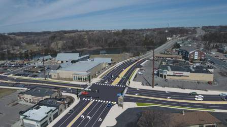 Aerial view of a busy intersection with multiple lanes, surrounded by commercial buildings, parking lots, and trees. Cars are visible on the roads, and a clear blue sky is in the background.