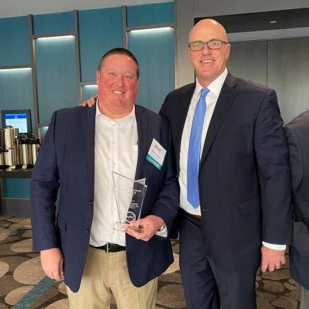 Two men in suits are standing in a conference room. The man on the left is holding a glass award and smiling. The man on the right, wearing glasses and a blue tie, is also smiling with his arm around the other mans shoulder.