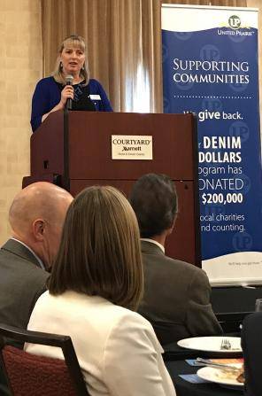A woman stands at a podium addressing an audience. Behind her is a banner reading “Supporting Communities” and “Denim Dollars” indicating a donation program. Several seated individuals listen attentively in the foreground.