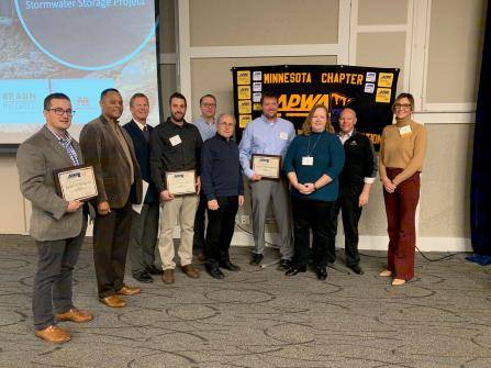A group of people stand together indoors, some holding certificates. They are in front of a banner that reads Minnesota Chapter and APWA. The setting appears to be an event or award ceremony.