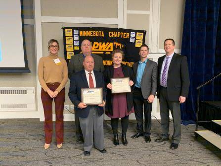 Six people are standing together indoors, holding certificates. They are in front of a banner that says Minnesota Chapter. The setting appears to be a formal event or presentation.