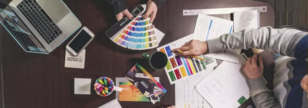 Aerial view of a cluttered office desk at a professional services firm with three people working. Items include a laptop, smartphone, colorful swatches, sketches, notes, and a coffee mug. Hands point to papers and hold swatches, indicating collaboration and strategic planning.