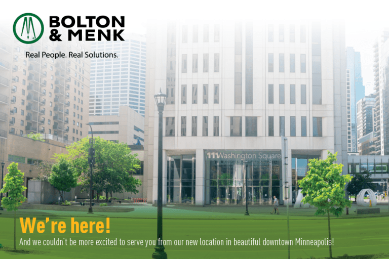 Front view of a tall building with large windows and a sign reading 111 Washington Square. The foreground features a grass lawn and trees. Text reads: Were here! And we couldnt be more excited to serve you from our new location in beautiful downtown Minneapolis! Logo upper left says Bolton & Menk.