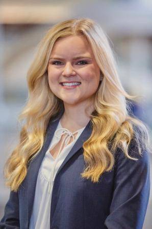 Smiling person with long, wavy blonde hair wearing a navy blazer over a white blouse, standing indoors with a blurred background.