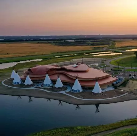 Aerial view of a unique building shaped like an abstract bird beside a river at sunset. The structure has a large, swooping roof and is surrounded by several white cone-shaped structures resembling tipis. Surrounded by fields and patches of water.